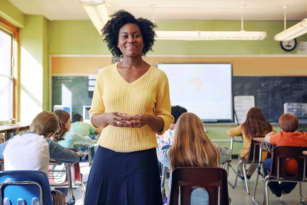 Teacher in yellow sweater in front of classroom with blackboard in background